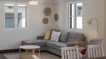 A modern living room with a grey sectional sofa, a round wooden table, and a white armchair. There are three windows with white frames, and a floor lamp is visible.