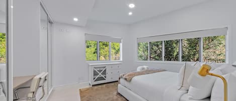 Een moderne slaapkamer met een groot bed, een bureau en een stoel. Er zijn meerdere ramen die natuurlijk licht en uitzicht op groen bieden.