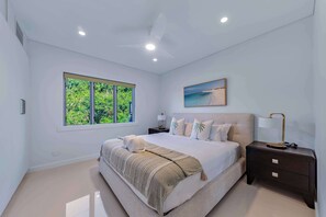 A modern bedroom with a large bed, a ceiling fan, a window with a view of greenery, and a painting of a beach. - Airlie Views Luxe Escape  - Ocean Views and Pool (Airlie Beach)