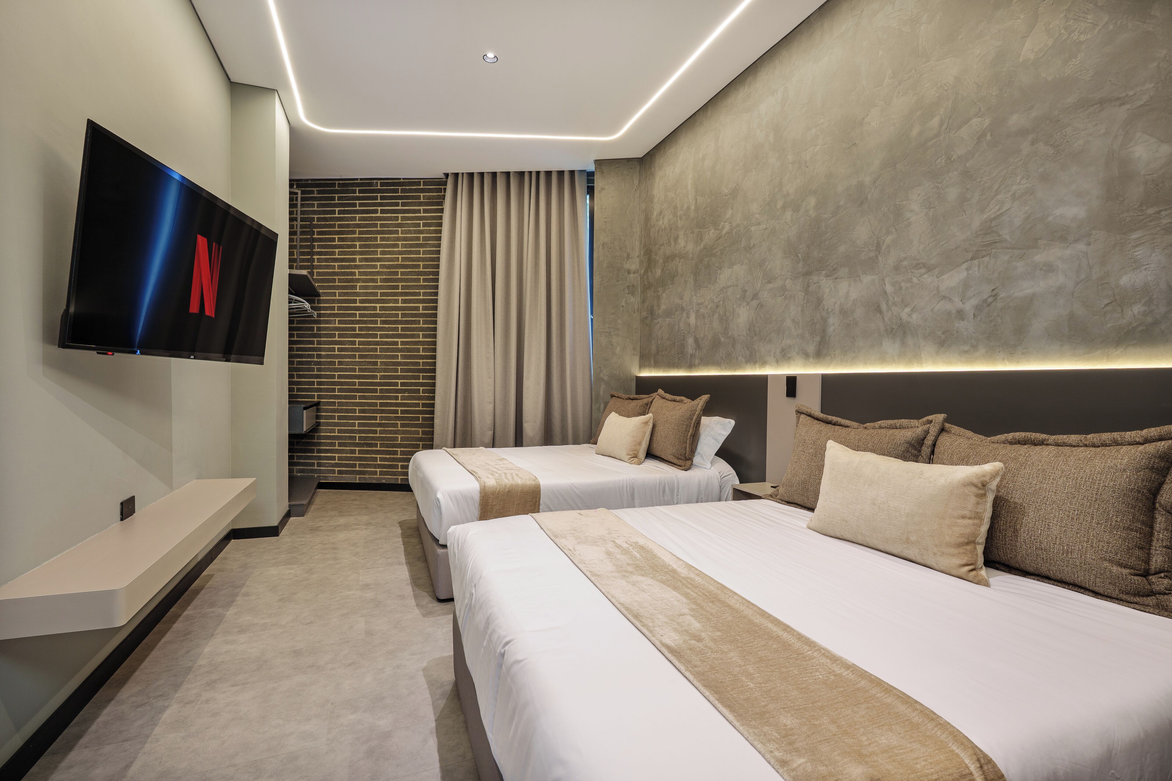 Quarto de hotel com duas camas, televisão de tela plana, parede de tijolos e design moderno no teto.