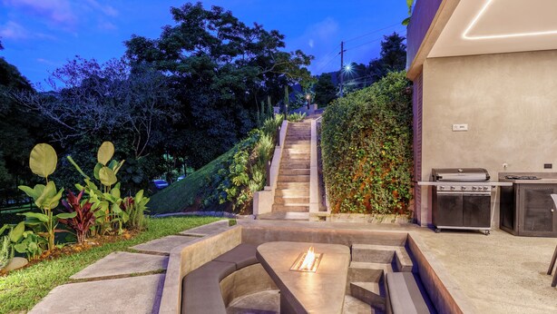 A modern outdoor patio with a built-in fire feature, a barbecue grill, and a staircase leading to a covered area.
