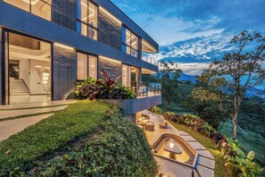 Una casa moderna de dos plantas con un balcón grande, mobiliario exterior y una chimenea.