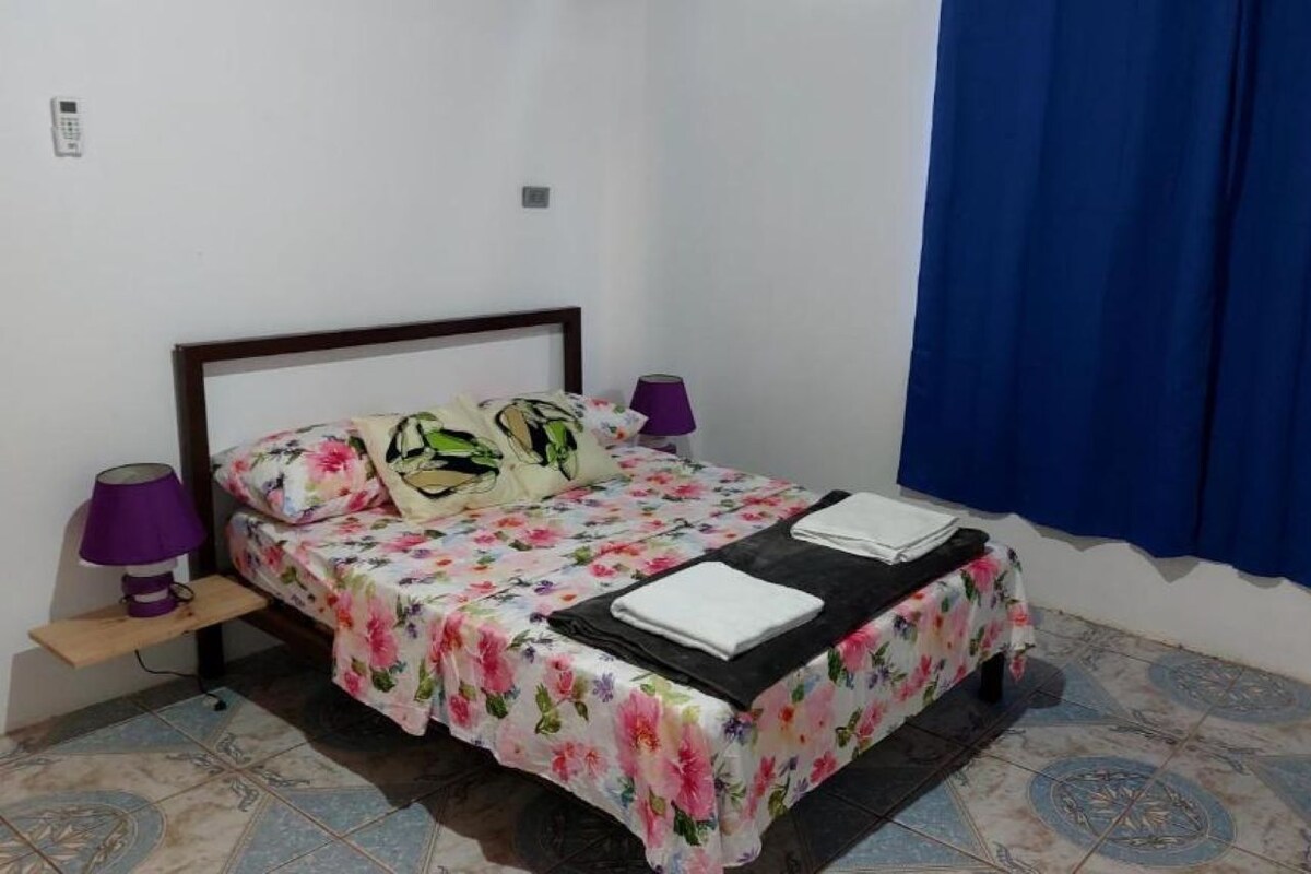 Un dormitori amb un llit, una tauleta de nit, una làmpada, una finestra amb cortines i un sòl de rajola.