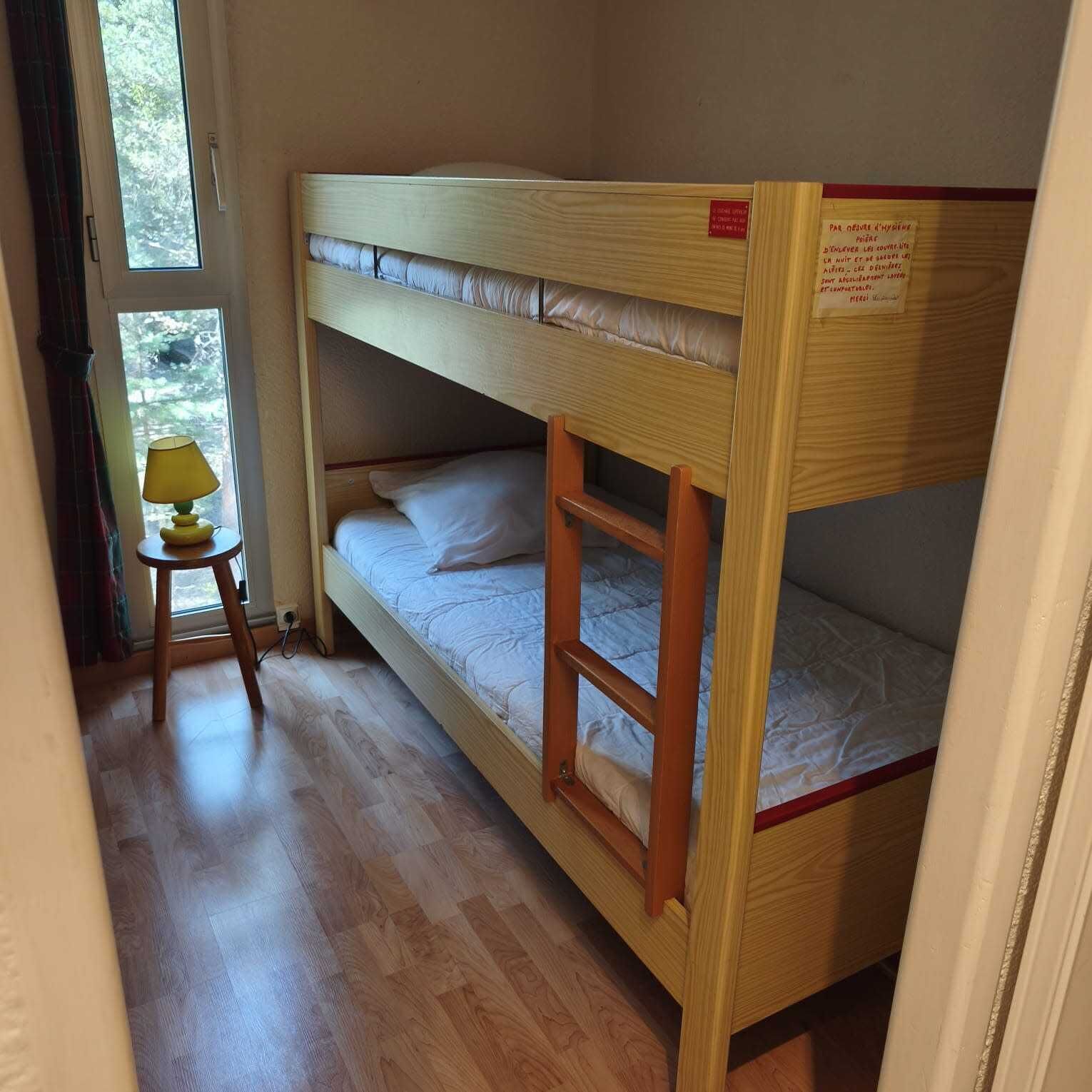 A bunk bed with a small table and lamp, wooden flooring, and a window with a view of trees.
