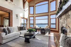 A spacious living room with a large window overlooking a body of water, a fireplace, and a cozy seating area. - Sandy Beach Lodge | Lakefront Luxury | 30min to YNP (Island Park)