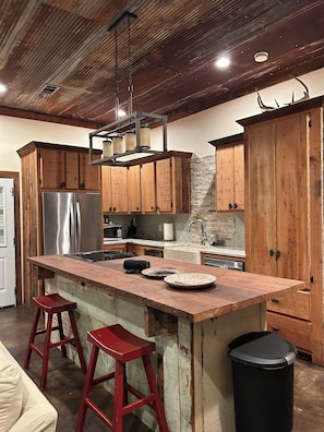 A kitchen with wooden cabinets, a stainless steel refrigerator, a wooden island with stools, and a black trash can.