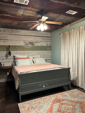 A bedroom with a bed, a ceiling fan, a window with curtains, a wooden wall, and a patterned rug.