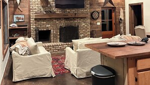 A rustic interior with a brick fireplace, white cushioned seating, a wooden ceiling, and a television mounted above the fireplace.