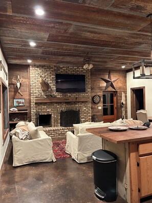 A rustic interior with a brick fireplace, white cushioned seating, a wooden ceiling, and a television mounted above the fireplace.