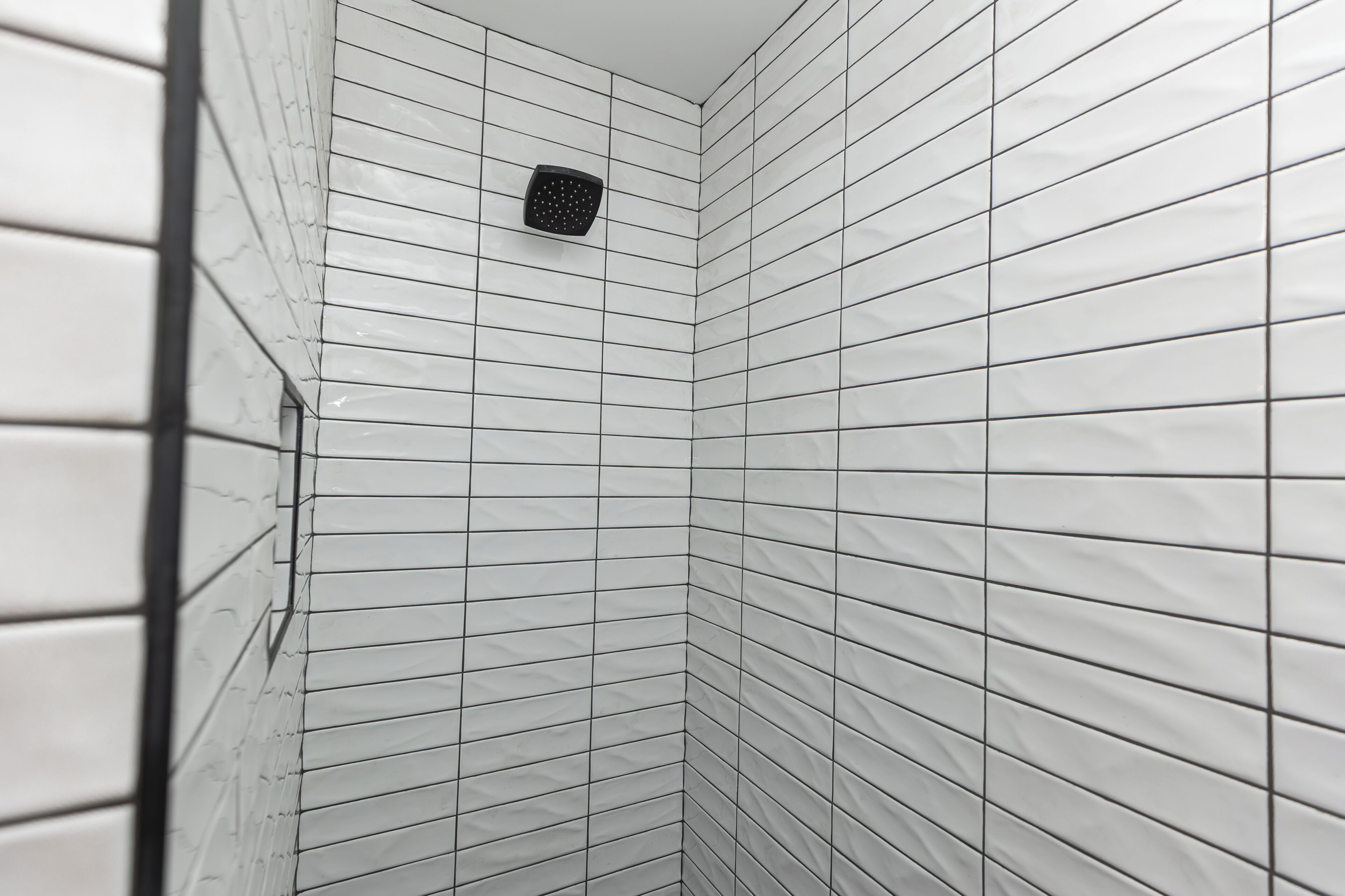 A corner of a room with white tiled walls and a black showerhead.