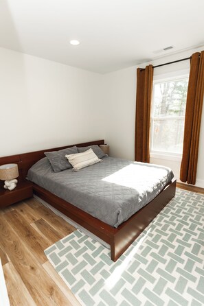 Ein modernes Schlafzimmer mit einem großen Bett, einem Holz-Kopfstück, einem Nachttisch mit Lampe und einem Fenster mit Vorhängen.