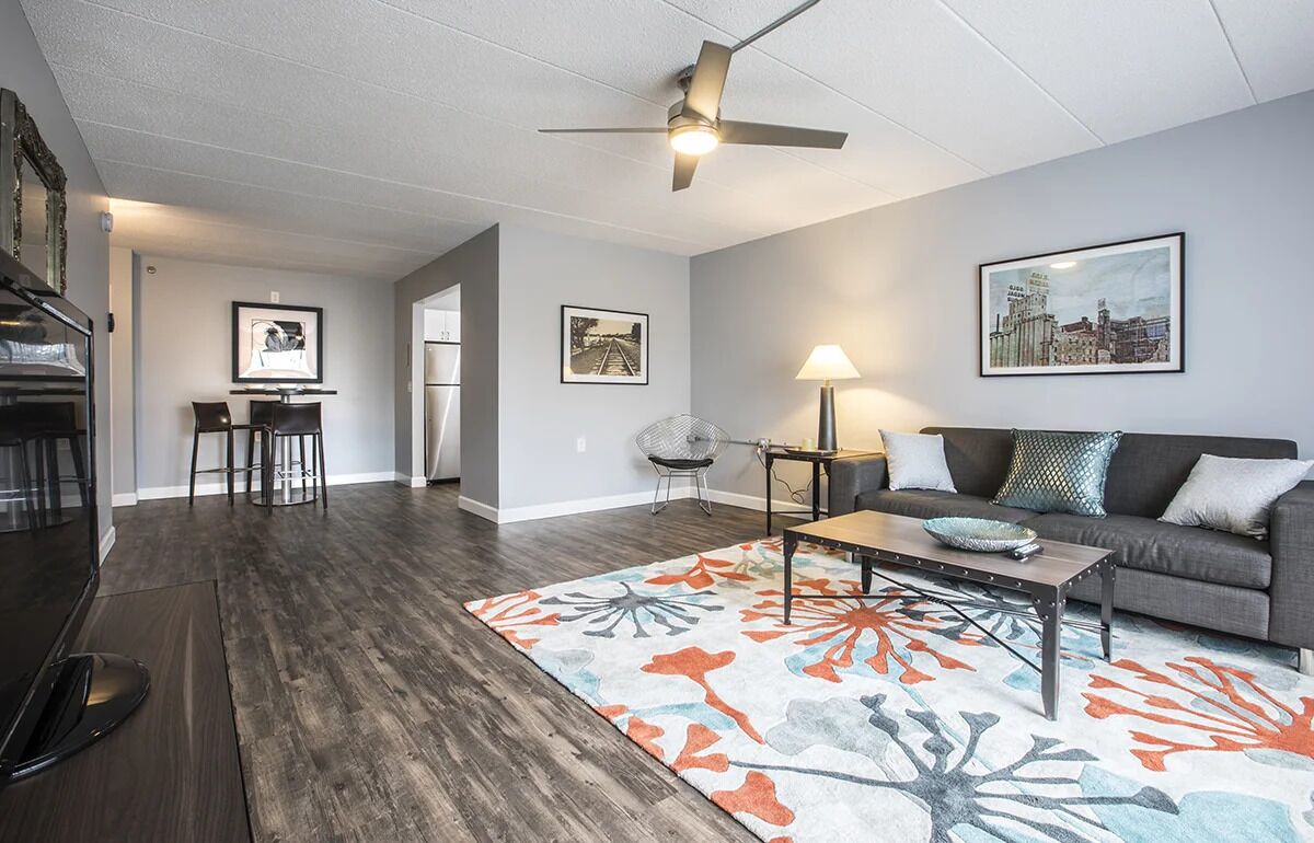 A modern living room with a sofa, coffee table, and a patterned rug. There is a dining area with chairs and a table, and a ceiling fan.