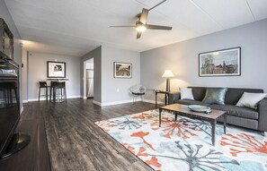 Ein modernes Wohnzimmer mit einer Couch, einem Couchtisch und einem gemusterten Teppich. Es gibt einen Essbereich mit Stühlen und einem Tisch, sowie einen Deckenventilator.