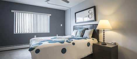 A bedroom with a bed, a nightstand, a lamp, a ceiling fan, and a window with blinds.