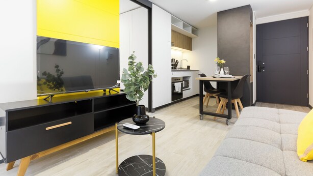 Ein modernes Wohnzimmer mit Flachbildfernseher, einem kleinen runden Tisch mit einer Vase, einer Couch und einer Küche im Hintergrund.