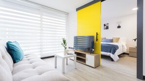 Ein modernes Wohnzimmer mit Sofa, Couchtisch, Fernseher und Bett, dessen Wand durch einen gelben Farbakzent hervorgehoben wird.