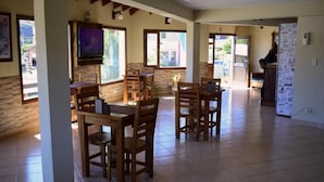 Un grand espace de salle à manger avec des tables et des chaises en bois, un téléviseur fixé au mur et un réfrigérateur décoré d’aimants.