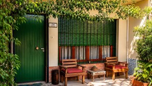 Une porte verte avec une pancarte, des bancs en bois avec des coussins rouges, une petite table et des plantes en pot.