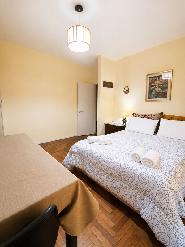 A hotel room with a bed, a table, a chair, a nightstand, a lamp, and a painting on the wall.