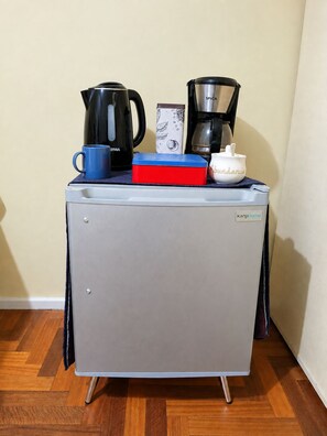 A compact kitchenette with a refrigerator, a coffee maker, a kettle, and a mug.