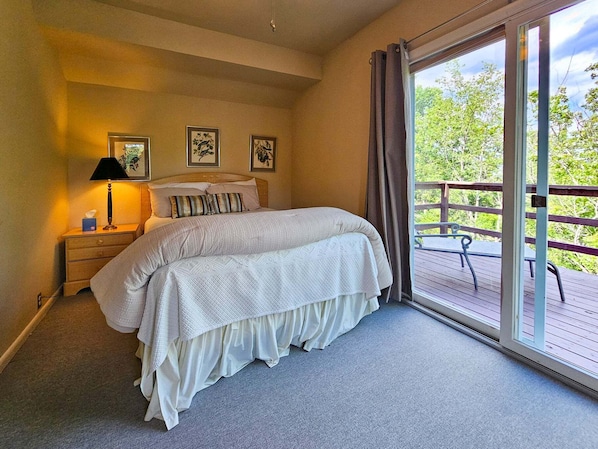 Quarto com cama, mesa de cabeceira, abajur e janela com vista para árvores.