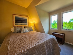 Quarto com cama grande, mesa de cabeceira, escrivaninha, janela com vista para vegetação e um quadro na parede.