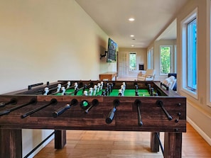 Uma mesa de futebol de botão em um ambiente com grandes janelas, uma televisão na parede e vista para o verde lá fora.