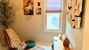Una habitación con una silla de madera, un cojín verde, una mesita con un jarrón y una ventana que da a un edificio.