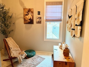 Una habitación con una silla de madera, un cojín verde, una mesita con un jarrón y una ventana que da a un edificio.