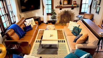 A living room with a brown leather sofa, a wooden coffee table, and a blue armchair. There is a television mounted on the wall and a fireplace with a decorative mantle.