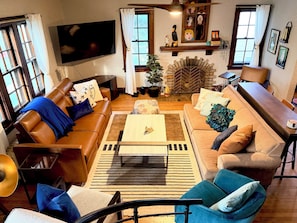 A living room with a brown leather sofa, a wooden coffee table, and a blue armchair. There is a television mounted on the wall and a fireplace with a decorative mantle.