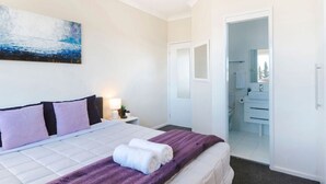 A hotel room with a bed, a nightstand with a lamp, a painting on the wall, and a bathroom visible through an open door. - Oceans 1, Beach Views, 3-Min Walk to Birubi Beach (Anna Bay)