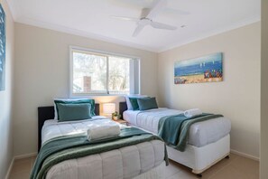 A bedroom with two beds, a ceiling fan, a window with a view, and a beach-themed painting on the wall. - Oceans 1, Beach Views, 3-Min Walk to Birubi Beach (Anna Bay)