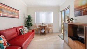 A living room with a red sofa, a dining table with chairs, a TV stand, and a large window with blinds. - Oceans 1, Beach Views, 3-Min Walk to Birubi Beach (Anna Bay)