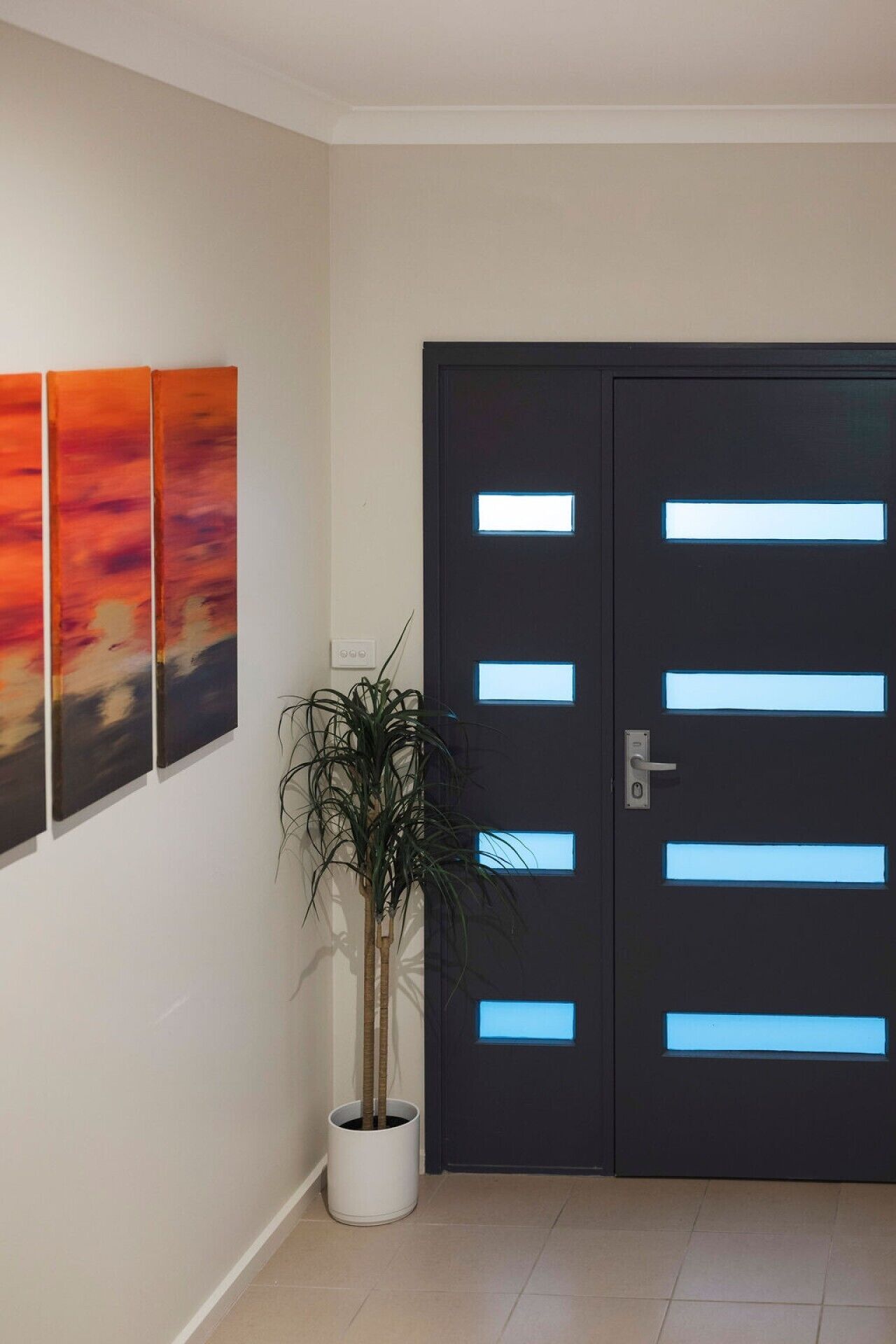 Un interior moderno con una puerta oscura que tiene paneles iluminados, una planta en maceta y un cuadro abstracto en la pared.