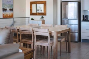 A dining area with a wooden table and chairs, a kitchen with a refrigerator, and a painting on the wall. - Oceans 2, Ocean Views, 3-Min Walk to Birubi Beach (Anna Bay)