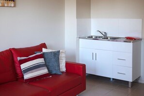 A compact living space with a red sofa, a kitchenette with a stainless steel sink, and white cabinets. - Oceans 2, Ocean Views, 3-Min Walk to Birubi Beach (Anna Bay)