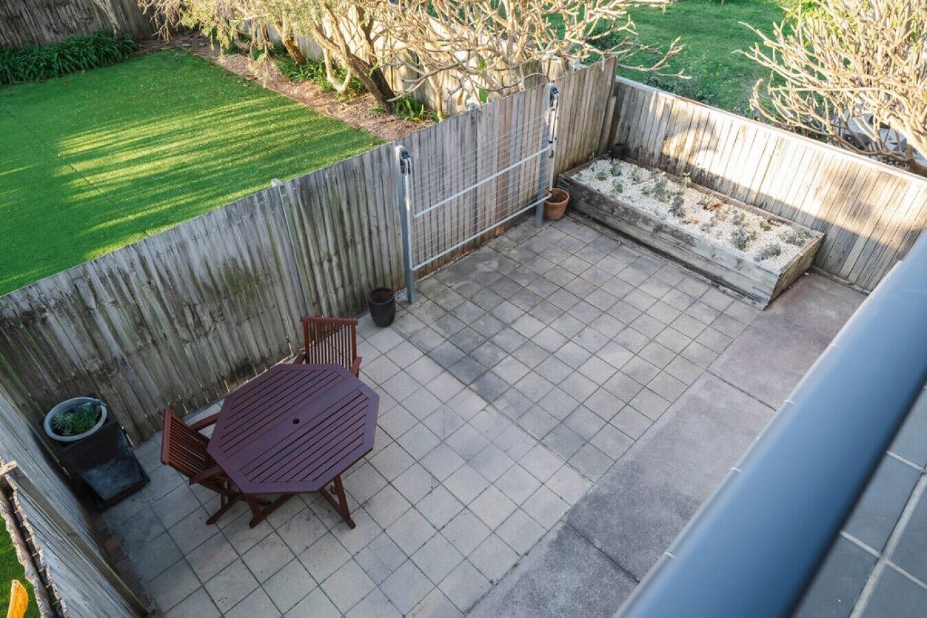 An outdoor patio with a tiled floor, a wooden table and chairs, a built-in bench, and a fenced area with a grassy lawn and trees.
