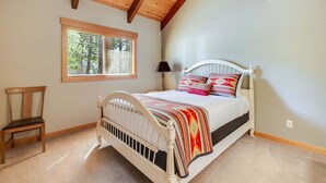 Ein Schlafzimmer mit Holzbalkendecke, einem Fenster mit Jalousien, einem Bett mit weißem Kopfteil und einem Sessel.
