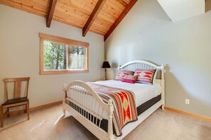 Ein Schlafzimmer mit Holzbalkendecke, einem Fenster mit Jalousien, einem Bett mit weißem Kopfteil und einem Sessel.