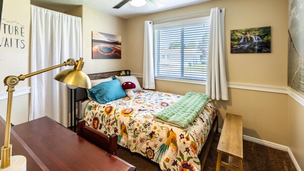 A bedroom with a bed, a desk, a chair, a ceiling fan, a window with blinds, and a wall with a painting. - Sunshine House | Bright & Cozy Getaway for Families, Friends, and Relaxation (Rexburg)