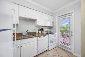 A kitchen with white cabinets, a granite countertop, a built-in microwave, a dishwasher, and a window with blinds. - 👣Barefoot Beach Resort #E211 - 2 Bedroom condo in Indian Shores Beach with a Pool!🩴 (Indian Shores)