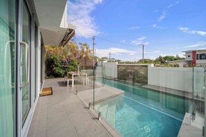 A modern outdoor swimming pool with clear water, surrounded by a glass enclosure and a stone wall. - Alex Headland Haven - 3 Level Modern Retreat (Mooloolaba Region)