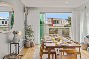 Een eethoek met een tafel voor vier personen, een spiegel, een potplant en een uitzicht op een huis door een glazen deur.