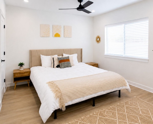 Un dormitorio con cama, mesitas de noche, ventilador de techo, ventana con persianas y decoración en la pared.
