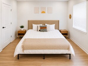 Un dormitorio con una cama, dos mesitas de noche, un ventanal con persianas y una puerta.