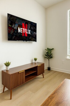 A modern living room with a flat-screen TV mounted on the wall displaying the Netflix logo, a wooden entertainment unit with two potted plants, and a large window with blinds. - Homey Worker-Friendly 2BR I Pool & Gym (Sherman)