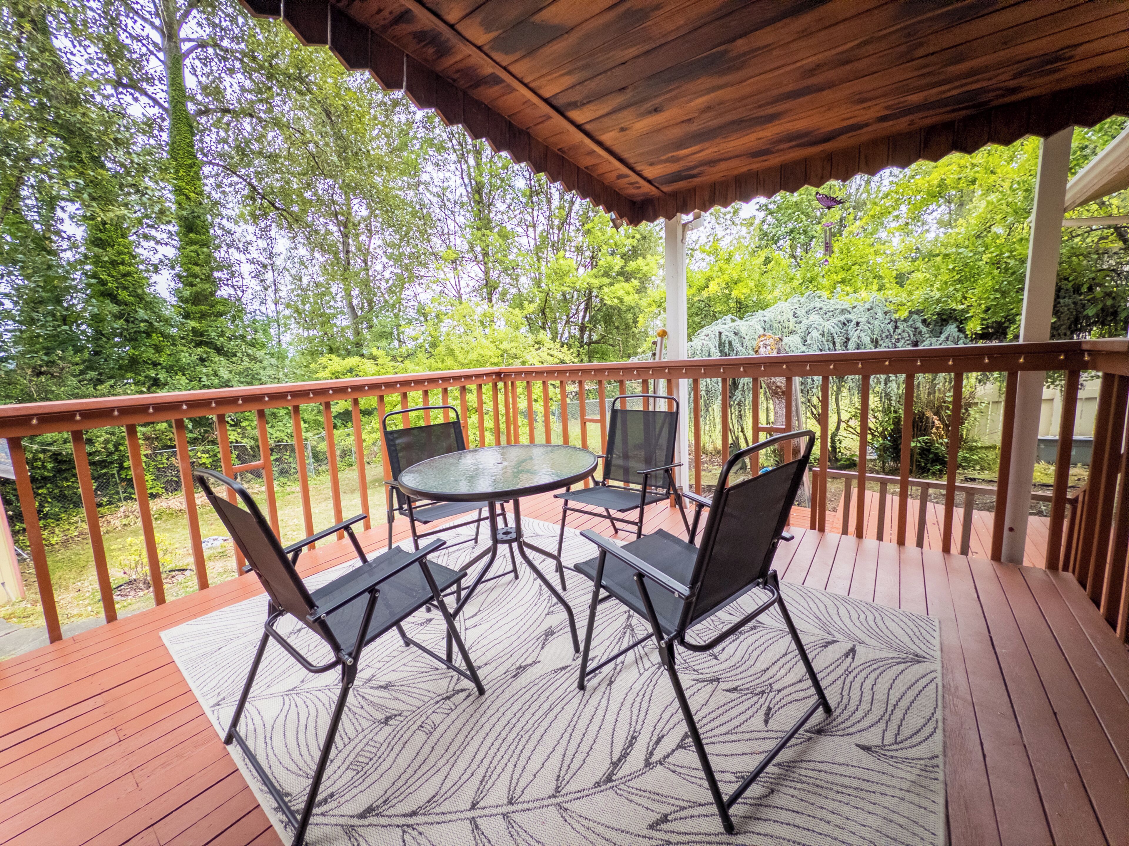 A covered outdoor deck with a table and chairs, surrounded by a wooden railing and greenery.