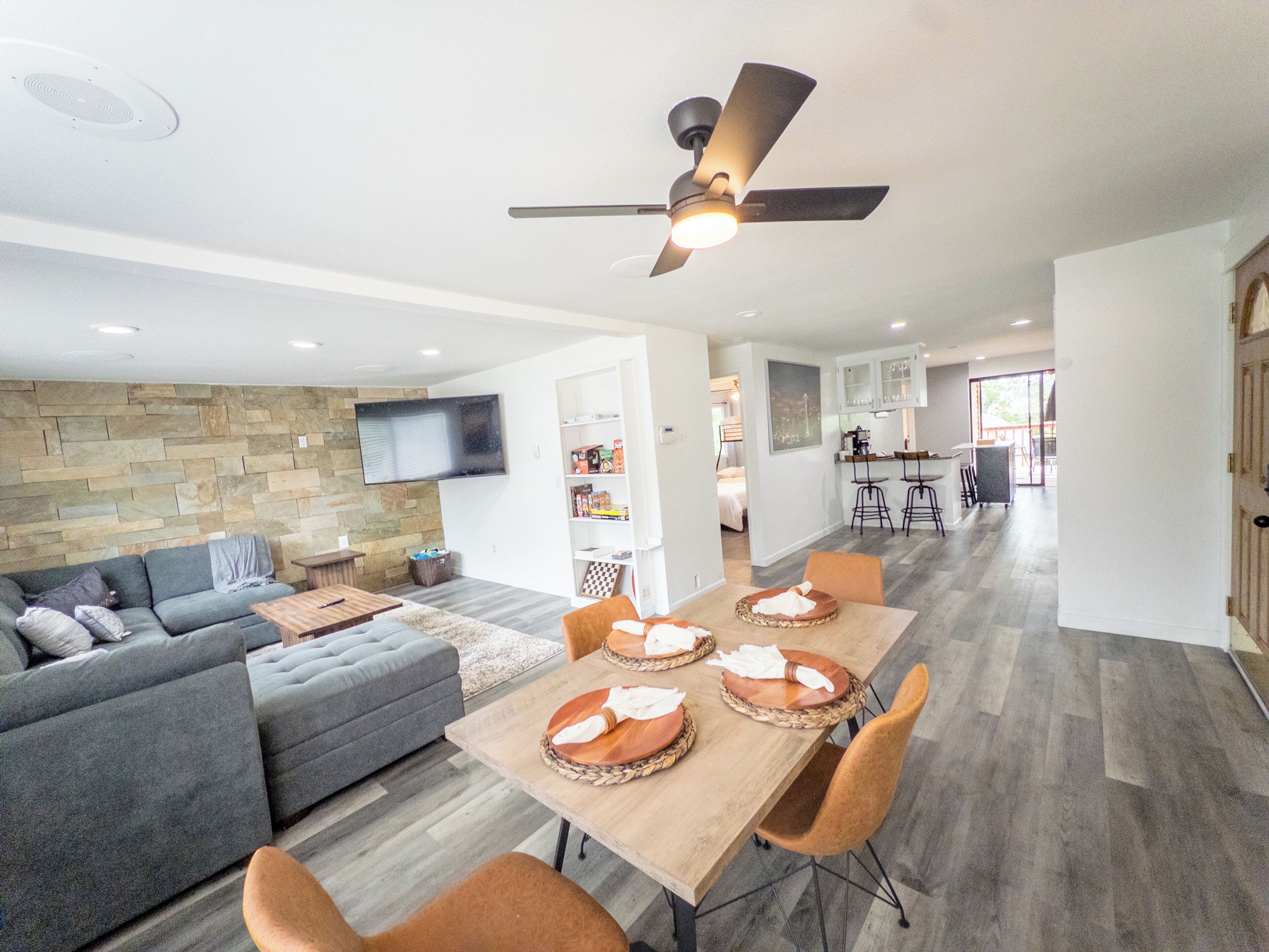 A modern living space with a dining area, a sofa, a television mounted on the wall, and a ceiling fan.