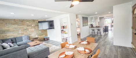 A modern living space with a dining area, a sofa, a television mounted on the wall, and a ceiling fan.